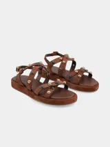 Sandalias planas de cuero gamuza color marrón chocolate, con tiras cruzadas decoradas con apliques metálicos y piedras. Cierre ajustable con hebilla en el tobillo y suela forrada.