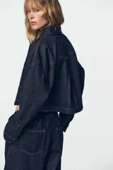 Camisa de denim color azul oscuro, con cuello solapa, manga larga y corte cropped. Presenta dos bolsillos de plastrón con tapa en el frente y cierre frontal con botones metálicos.