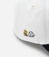 Gorra blanca de sarga con visera azul marino y bordado de Snoopy jugando baseball.