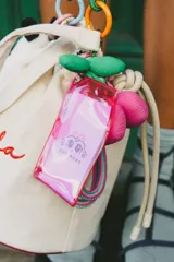 Llavero multifuncional con tarjetero de acrílico fucsia translúcido, decorado con un dije de cerezas de tela en color fucsia y verde, y una correa de cuerda trenzada en tonos fucsia y verde con detalles en lila.