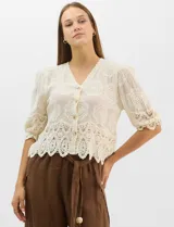 Blusa de algodón semitransparente con diseño floral bordado a tono. Presenta escote en V, cierre frontal con botones y mangas al codo ligeramente abullonadas. Incluye detalles de encaje calado estilo macramé en los puños y en el ruedo con terminaciones en ondas.