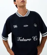 Remera de tejido texturizado color azul marino, con cuello redondo y mangas cortas con puños a rayas blancas. Presenta bordados frontales con la inscripción 'Future Co.', el número '98', un logo ovalado y estrellas.