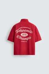 Camisa de algodón con estructura canvas, color rojo lavado, de corte relajado (relaxed fit) y manga corta. Posee cuello solapa, dos bolsillos de plastrón en el pecho y cierre frontal de botonadura. Presenta bordado y estampado de texto a contraste en el delantero.