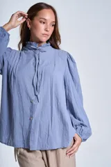 Camisa de color azul con cuello alto decorado con volados y lazo de ajuste. Presenta tablas decorativas en los hombros, cierre frontal con botones de nácar y mangas largas con puños elásticos que generan un efecto abullonado.