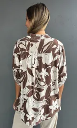 Blusa de corte holgado con estampado floral en tonos marrón y blanco. Presenta cuello camisero, cierre frontal con botones y un bolsillo en el pecho con detalle de lentejuelas. Las mangas son tres cuartos con trabas para ajustar.