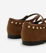 Zapatilla tipo Mary Jane color marrón con efecto gamuzado, tira fina con hebilla ajustable sobre el empeine y pequeñas tachuelas metálicas doradas. Tiene punta redonda y suela baja.