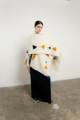 Ruana de tejido grueso color crema con flecos en el borde inferior y diseño geométrico en ocre, negro y gris.