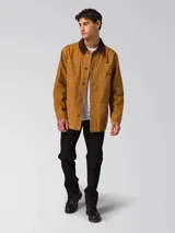 Chaqueta tipo chore de lona color marrón, con cuello camisero en contraste color marrón oscuro, cierre frontal con botones, cuatro bolsillos de parche frontales y costuras a la vista en color blanco.