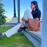 Cartera tipo morral en eco cuero con textura símil gamuza marrón, costuras blancas visibles y correa ajustable de lona.