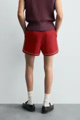 Short de algodón color rojo oscuro, de corte regular fit, con cintura ajustable mediante cordón blanco. Presenta ribetes o vivos de contraste en color blanco bordeando los laterales, el bajo y los bolsillos.
