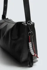 Bolso tipo bandolera de diseño minimalista en color negro, con cierre de cremallera superior y asa de hombro ajustable. Incluye un detalle decorativo de charms metálicos en uno de los laterales.
