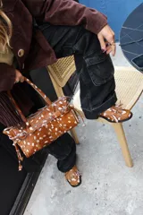 Cartera de tamaño medio confeccionada en cuero vacuno con textura de pelo y estampado animal print tipo vaca. Presenta estructura rígida, doble asa de hombro, cierre superior, detalle de tira horizontal con tachas y hebillas metálicas, y lazos decorativos en los laterales.