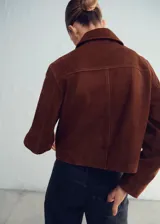 Chaqueta corta de piel de cabra color marrón, con solapas clásicas, cierre frontal de botones y bolsillos de parche.