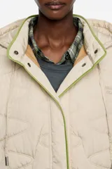 Chaquetón corto de corte línea A, confeccionado en tejido técnico acolchado color beige claro, con costuras en patrón geométrico. Posee cuello subido con capucha integrada, cierre frontal oculto con botones de presión, manga larga con puño elástico y vivos de ribete verde lima al contraste.