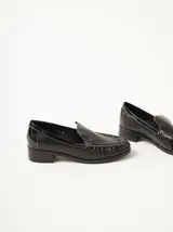 Mocasines de cuero negro con costuras pespunteadas en hilo blanco a contraste, diseño fruncido en la puntera, talón plano con pequeño tacón y apliques sutiles en charol.