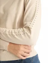 Buzo tejido color beige de manga japonesa, con detalle de tachas metálicas a lo largo de las mangas. Presenta puños y ruedo acanalados.