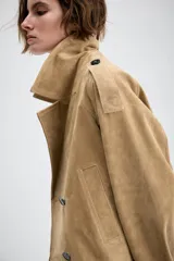 Trench corto cruzado de piel de ante color tostado, con cuello de solapa, bolsillos delanteros de vivo y manga larga con trabillas en los puños.