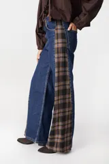 Jean de pierna ancha con diseño bicolor, combinando denim azul clásico con un panel lateral de tela a cuadros en tonos marrón y negro. Presenta cuatro bolsillos funcionales y cierre frontal.