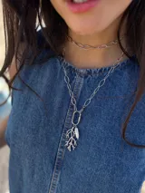 Collar largo de acero quirúrgico plateado con cadena de eslabones ovalados. Presenta un dije en forma de rama de coral y una perla de río natural, unidos a través de un mosquetón ovalado funcional que permite desmontar los dijes o ajustar el largo del collar.