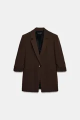 Blazer negro con cuello solapa con muescas, hombreras y forro interior a tono. Presenta manga por debajo del codo con detalle de drapeado y falsos bolsillos de vivo en el delantero, con cierre frontal de un solo botón dorado.