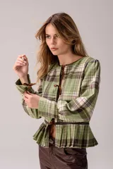 Blusa de tartán en tonos verdes y marrones, con detalles en cuero de oveja. Presenta un diseño versátil con cierre frontal mediante tiras de cuero para anudar, mangas largas con puños ajustables y un ruedo con tablas que le otorga volumen. Puede utilizarse cerrada, abierta como saquito o invertida.