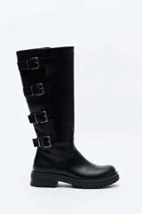 Bota alta de cuero sintético negro con suela track y tacón bajo. Presenta detalle de cuatro correas con hebillas metálicas en el lateral exterior de la caña.