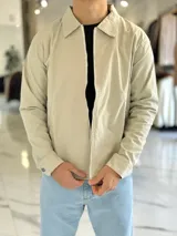 Campera de gabardina beige de corte boxy fit, con cuello camisero, cierre frontal de cremallera blanca y bolsillos laterales ribeteados.