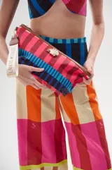 Necessaire de lona estampada con diseño de rayas multicolor. Presenta detalles en cuero ecológico, cierre superior metálico, aplique decorativo en forma de caracola dorada y correa lateral para llevar en la mano.