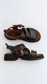 Sandalias de cuero marrón oscuro con efecto desgastado, tiras cruzadas en el empeine, hebilla metálica lateral y suela track negra con plataforma.