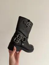 Bota de caña media en eco cuero negro, con diseño estilo biker que incluye correas con hebillas metálicas y herrajes plateados en el tobillo. Cuenta con suela plana y punta redondeada.