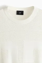 Remera de malla color blanco hueso, de manga corta, con cuello redondo y hombros caídos. Presenta un diseño de tejido perforado con un número grande grabado en el frente.