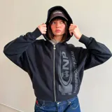 Campera tipo hoodie con cierre frontal, color negro, de corte oversize. Presenta un estampado gráfico en tono gris oscuro en el frente con el logo de la marca y la inscripción Nude Project Team, además de un pequeño bordado de una cereza en la manga.