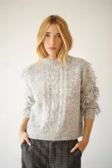 Buzo de punto color gris melange, con cuello redondo, mangas largas con flecos y diseño de trenzas en el frente.