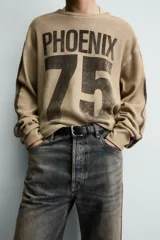 Sudadera de corte relajado confeccionada en tejido con textura waffle, color beige con efecto lavado. Presenta cuello redondo, manga larga con franjas laterales oscuras y estampado frontal con texto y número en contraste.