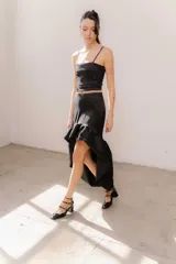 Pollera midi de tafeta color negro, con diseño asimétrico de corte mullet (más corta adelante y larga atrás), tiro alto y detalle de volado fruncido en el ruedo.