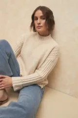 Polera de tejido de punto color beige, con cuello alto, diseño de ochos en los hombros y mangas largas con puños doblados.