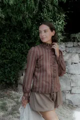 Camisa de manga larga con estampado de rayas verticales en tonos marrones y rojizos. Presenta cuello alto con volados, cierre frontal con botones y detalle de volado lateral a lo largo de la botonadura. Las mangas cuentan con puños elásticos con volados.