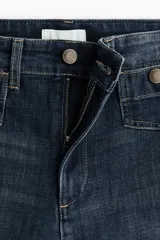 Jean tobillero de denim azul con leve elasticidad, tiro alto y perneras amplias tipo wide leg. Presenta bolsillos delanteros tipo parche con solapa y botones metálicos, además de pliegues marcados al frente y en la espalda.