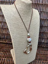 Colgante con cordón de cuero marrón, decorado con cuentas de piedra en tonos blanco y marrón, y un dije metálico dorado de forma irregular.