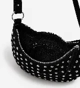 Bolso de hombro estilo hobo en color marrón oscuro, confeccionado en gamuza con diseño de tachas metálicas plateadas distribuidas por toda la superficie. Cuenta con un asa superior fija y cierre superior con cremallera.