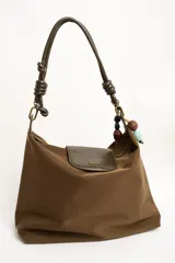 Cartera tipo shopper color marrón, con solapa frontal decorativa en tono más oscuro y tiras con nudos. Incluye herrajes dorados y un charm colgante con cuentas de madera.