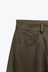 Pantalón de tiro medio color verde oliva, confeccionado en tejido con estructura de espiga. Presenta pierna ancha, trabillas en la cintura, cinco bolsillos y cierre frontal con cremallera y botón.