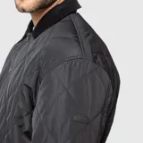 Chaqueta acolchada de diseño romboidal en color negro, con cuello camisero de textura aterciopelada y cierre frontal mediante botones a presión.