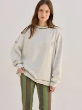 Buzo oversize color gris melange con corte crew, cuello redondo, mangas largas y detalles bordados en color verde en el cuello, puños y ruedo. Presenta el logo de la marca bordado en el pecho.