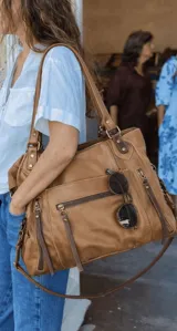 Cartera tipo tote bag de cuero color tostado, con diseño funcional que incluye múltiples bolsillos exteriores con cierre, herrajes metálicos y correa larga ajustable extraíble. Presenta un acabado suave y costuras reforzadas.