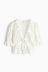 Blusa color blanco crudo, de escote en V pronunciado con ribetes de volado, mangas abullonadas al codo y detalles de alforzas, encaje y bordados en el frente.