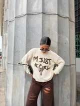 Sweater de punto color crema con cuello redondo y mangas largas. Presenta un estampado frontal en color marrón con la frase 'I'm just a girl' en estilo grafiti.