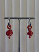 Aros colgantes compuestos por una argolla de acero quirúrgico y un dije de cristal de Murano en color rojo con forma de espiral retorcida.