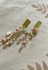 Aros colgantes con base rectangular dorada texturizada y múltiples cuentas de distintos tamaños y formas en tonos rosa, blanco y dorado, unidas con alambre dorado.