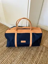 Bolso de viaje tipo duffel de lona negra con detalles en cuero tostado, asas de mano y correa ajustable para el hombro.